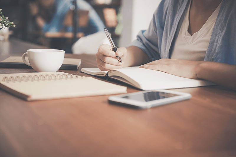 femme qui écrit sur un carnet à côté de son téléphone et de sa tasse de café